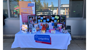 Freshers' Fair stall at West Lothian College. The table has a white table cloth with the Girlguiding Scotland logo at the front. There are colourful leaflets and information signs on a display board at the back of the table. To the back left there is a pull up banner with Girlguiding branding on it. 